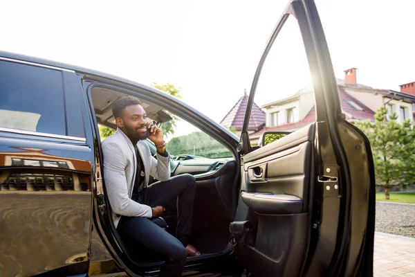 Joven hombre africano sonriente usando teléfono inteligente mientras está sentado en el asiento del pasajero en el coche. Concepto de gente de negocios — Foto de Stock