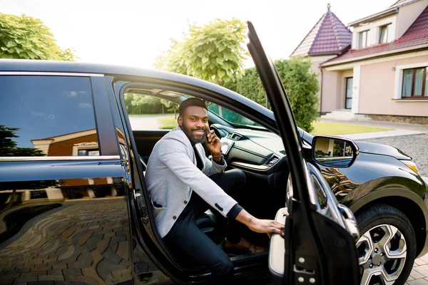 Heureux homme d'affaires afro-américain souriant en costume décontracté intelligent parlant sur smartphone tout en montant dans la voiture noire sur le siège passager . — Photo