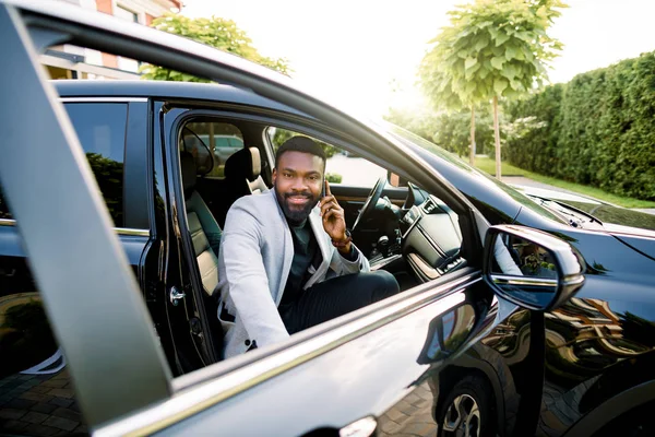 Happy smiling African american businessman in smart casual suit speaking on smartphone while getting in the black car on passenger seat. — 스톡 사진