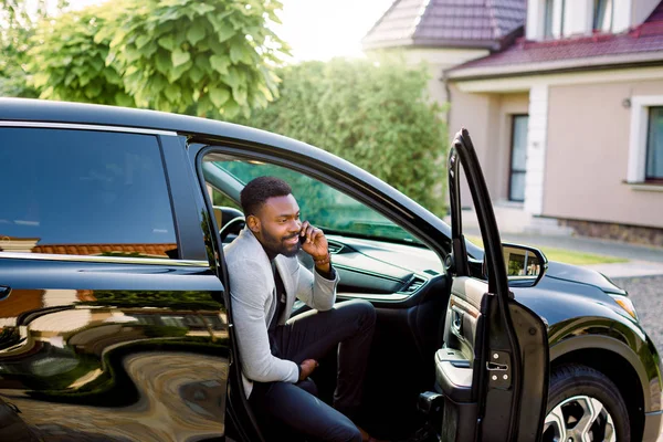 Pemuda tersenyum tampan Afrika pengusaha duduk di mobil di kursi penumpang dan berbicara di telepon. Bangunan, rumah dan pohon-pohon hijau dengan latar belakang teh — Stok Foto