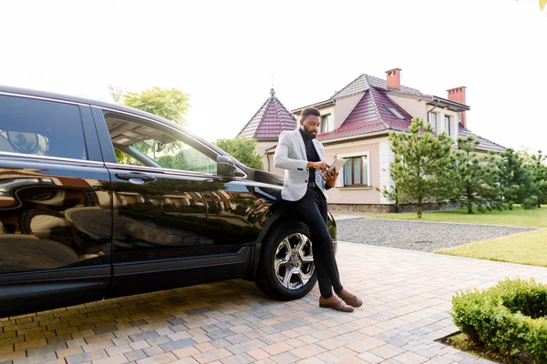 African american businessman in a suit waiting for meeting, scrolling news on gadget, or writing an email, while standing near his luxury black car crossover, outdoors, building on the background — 스톡 사진