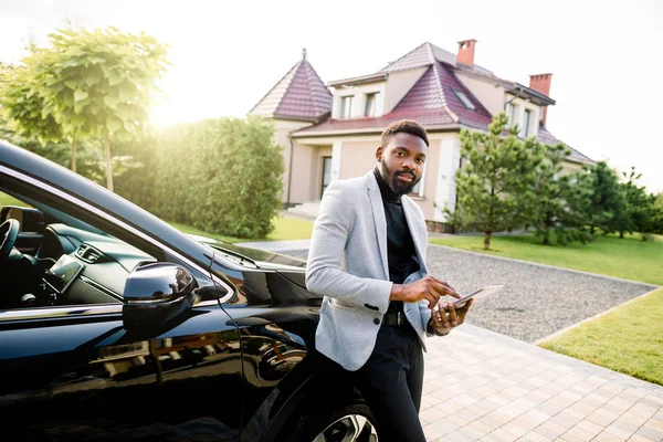 Beau Afro-Américain confiant tapant un e-mail sur smartphone, se tenant près de sa voiture de luxe, et regardant la caméra — Photo