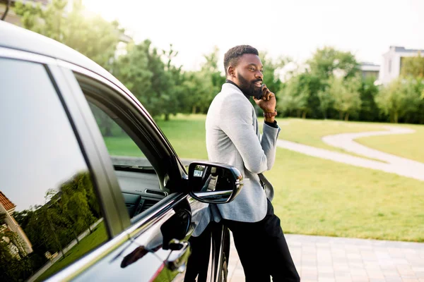 Homme d'affaires afro-américain en costume parlant sur smartphone tout en se tenant à l'extérieur près de sa voiture noire de luxe. Parc vert, arbres en arrière-plan — Photo