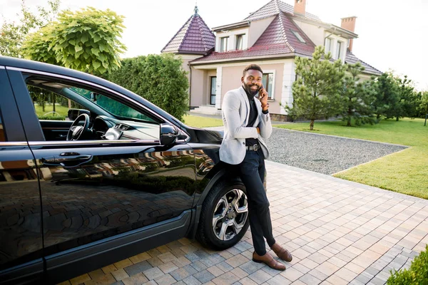 Handsome black businessman in a suit talking by smartphone near a black car, outdoors, building on the background
