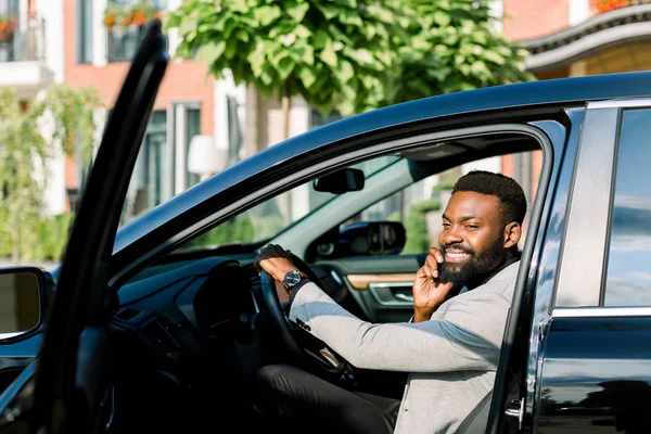 Joven empresario africano sentado en su coche negro y haciendo una llamada telefónica, y sonriendo. Edificios del centro de oficinas en el fondo — Foto de Stock