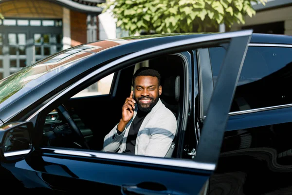 Pria Afrika ceria berjaket gey duduk di mobil berbicara dengan telepon, kantor pusat di latar belakang — Stok Foto