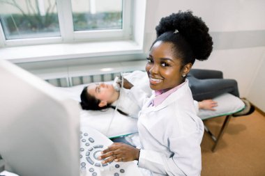 Ultrason sensörlü neşeli kadın Afrikalı doktor, kadın hastasına ultrason taraması yapıyor, modern hastane muayene odasında.