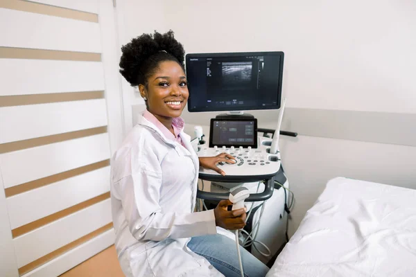 Smiling African woman doctor with ultrasound scanner in hand, working on modern ultrasound scanning machine in light room in clinic. Portrait Of 4D Ultrasound Scanning Machine Operator — 스톡 사진