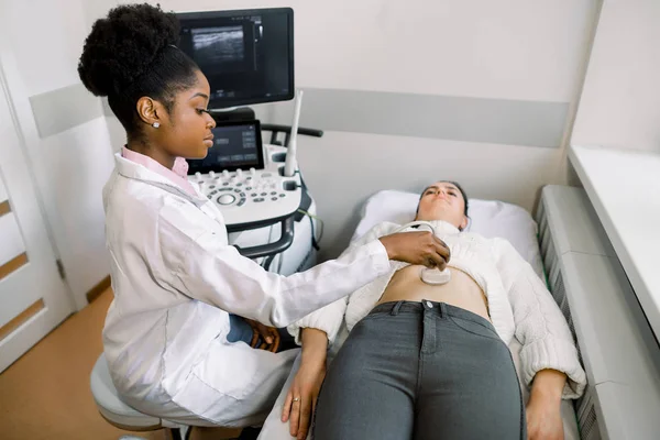 Mujer joven africana médico que mueve transductor de ultrasonido en el vientre de las mujeres en el hospital. Médico mujer dando paciente ultra sonido de abdomen. Mujer embarazada que se hace una prueba de ultrasonido en el consultorio del ginecólogo —  Fotos de Stock