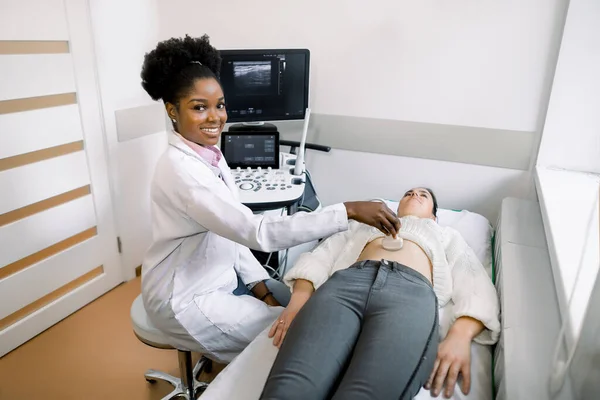 Молодая чернокожая женщина врач-сонограф с помощью ультразвукового аппарата на работе, держа в руке ультразвуковой сканер во время обследования женского живота пациента в больнице . — стоковое фото