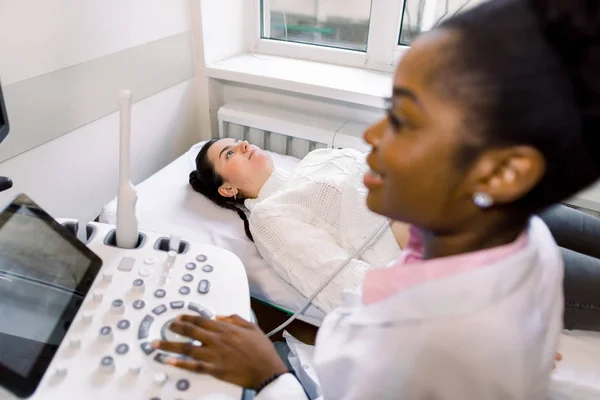 Медицинское обследование молодой белой женщины с помощью ультразвукового оборудования. Крупный план африканской женщины-врача, перевозящей ультразвуковой зонд на беременных женщинах Живот в больнице — стоковое фото