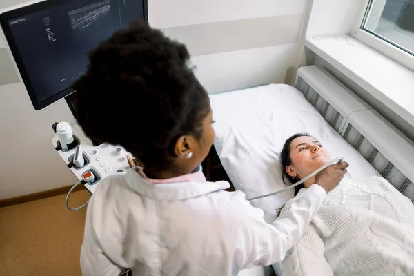 Young Caucasian woman patient during the ultrasound examination of a thyroid lying on the couch in medical office. African woman doctor and patient. Ultrasound equipment — Stock Photo, Image