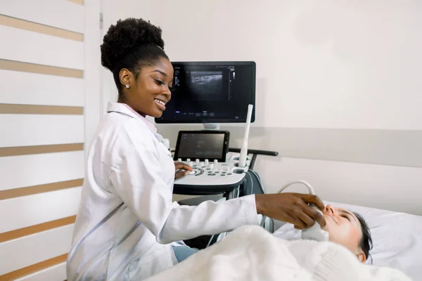 Young African female doctor using sonogram on neck of female patient in examination room