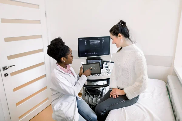 African female doctor gynecologist showing echo ultrasound photo on digital tablet to happy pregnant woman in modern medical office — Stock Photo, Image