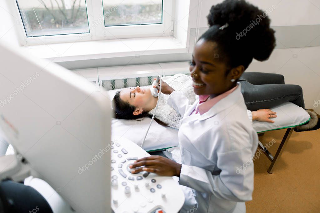 Attractive young woman having thyroid ultrasound scanning test by her female African doctor at the modern clinic