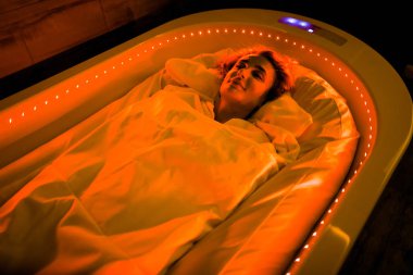 Spa vücut sargıları. Kaplıcada, sıcak yatak örtüsüyle sarılı, sıcak kapsülde kırmızı ışıkta uzanan, vücuduna çamur bulaştıran genç bir kadının fotoğrafı.