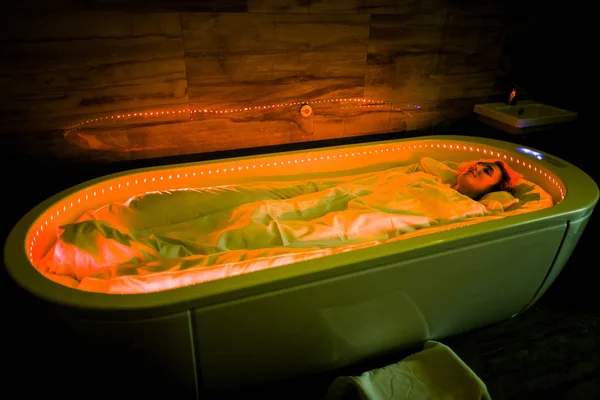 Envolturas corporales de spa. Foto de una mujer joven que se relaja en el salón de spa, envuelta con una cubierta de cama caliente especial, tumbada en una cápsula de spa caliente con luz roja, que tiene tratamiento de lodo del cuerpo —  Fotos de Stock