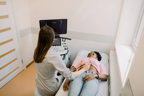 Young woman doctor looks at the ultrasound screen in a modern clinic, while performing ultrasound for young African pregnant woman — Stock Photo, Image