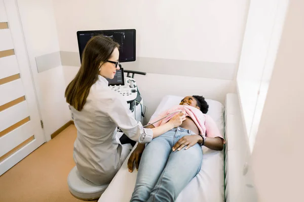 Happy young African female patient going through abdomen ultrasound at clinic. Professional Caucasian woman doctor doing stomach ultrasound — Stock Photo, Image