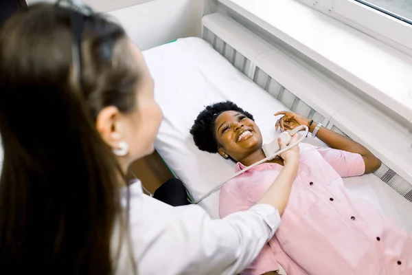 УЗИ шеи в клинике. Женщина-врач проводит исследования для молодой африканской улыбающейся пациентки . — стоковое фото
