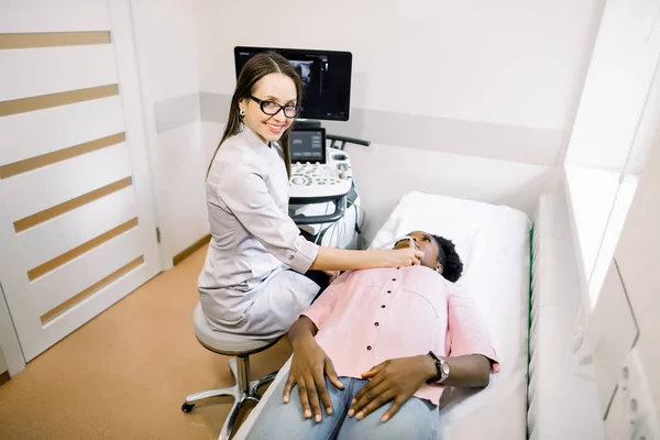 Молодая улыбающаяся женщина-врач в диагностическом центре делает УЗИ щитовидной железы для африканской пациентки. Женщина получает ультразвук щитовидной железы от врача — стоковое фото