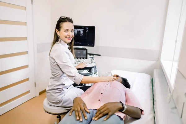 Médico de la mujer caucásica que examina la glándula tiroides de la mujer africana joven usando la máquina del escáner del ultrasonido. El médico pasa el sensor de ultrasonido sobre el cuello de los pacientes y mira a la cámara y sonríe —  Fotos de Stock