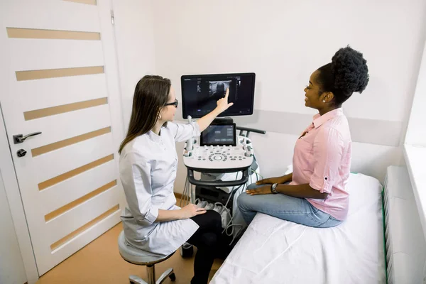 Fotografía de una hermosa joven africana sonriendo mientras estaba sentada en el sofá en la sala de examen ultrasonido y consultando a su médico mostrando los resultados de la prueba de ultrasonido en la pantalla —  Fotos de Stock