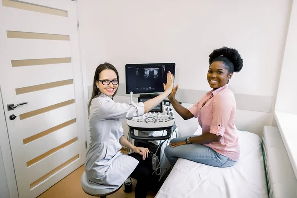 Gute Nachrichten nach Ultraschalldiagnostik. junge afrikanische Patientin, die sich glücklich und lächelnd fühlt, während sie ihrer lächelnden Ärztin vor laufender Kamera fünf gibt — Stockfoto