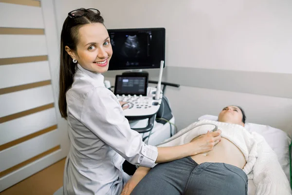 Молодая улыбающаяся женщина врач-сонограф с помощью ультразвукового аппарата во время проведения УЗИ брюшной полости пациентки в больнице — стоковое фото