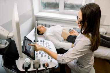 Genç bayan doktor kadın hasta için karın ultrasonuna hazırlanıyor. Doktor ultrason prosedürüne başlamadan önce kontrol panelindeki düğmelere basar.