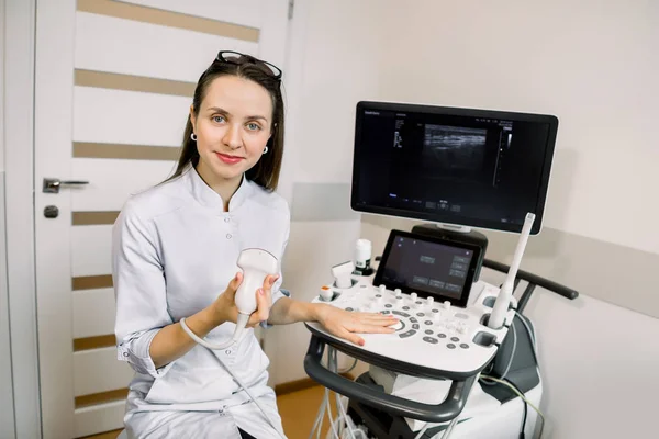 Profissional jovem médico ultrassonografista sentado nas proximidades moderna máquina de ultra-som e sorrindo — Fotografia de Stock