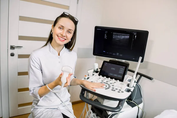 Profissional jovem médico ultrassonografista sentado nas proximidades moderna máquina de ultra-som e sorrindo — Fotografia de Stock