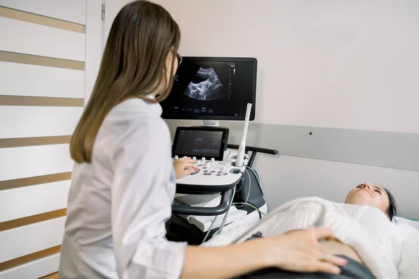 Mulher médico realiza um diagnóstico de ultra-som do abdômen paciente e órgãos internos, olhando para a tela da máquina de ultra-som moderna. Uma menina está em um sofá em uma sala de diagnóstico de ultra-som — Fotografia de Stock
