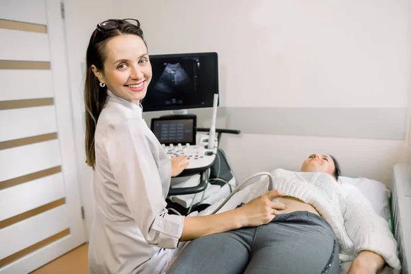 Jovem mulher sorridente médico ultrassonografista usando máquina de ultra-som durante a realização de teste de ultra-som do abdômen paciente feminino no hospital — Fotografia de Stock