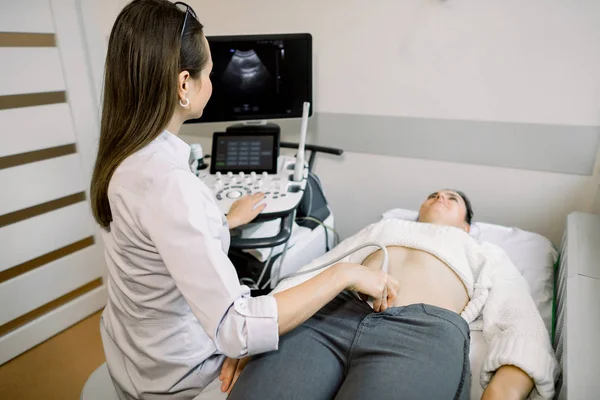 Mulher médico realiza um diagnóstico de ultra-som do abdômen paciente e órgãos internos, olhando para a tela da máquina de ultra-som moderna. Uma menina está em um sofá em uma sala de diagnóstico de ultra-som — Fotografia de Stock