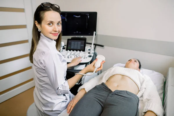 Счастливая женщина-врач держит ультразвуковой сканер и готова провести ультразвуковое сканирование молодой беременной женщины в современном медицинском родильном доме — стоковое фото