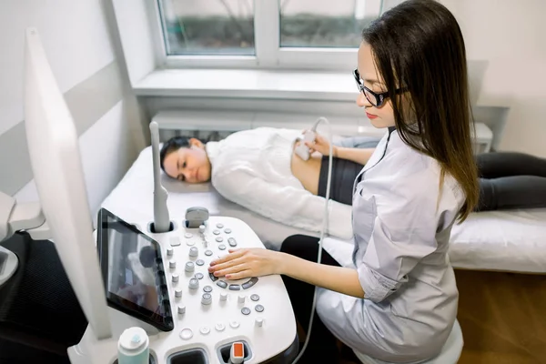 Professiona terapeuta sonógrafa que trabaja con la máquina de ultrasonido en la clínica, haciendo ultrasonido renal para la paciente femenina, mirando a la pantalla y pulsando botones en el panel de control —  Fotos de Stock