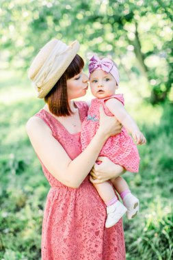 Hasır şapkalı güzel anne ve küçük kızı açık havada pembe bir elbise içinde. Mutlu bir ailenin açık hava portresi. Aile görünüşü. Anne kızını yanağından öpüyor..