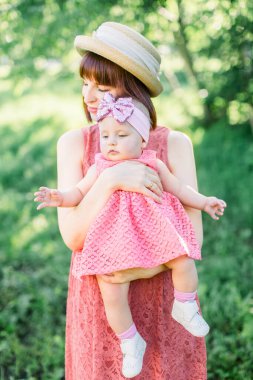 Hasır şapkalı güzel anne ve küçük kızı açık havada pembe bir elbise içinde. Çayırda küçük bebeğiyle genç bir anne.