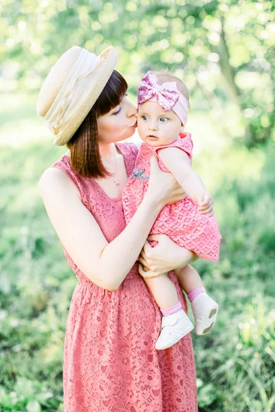 藁帽子の美しい母と彼女の小さな娘屋外の家族はピンクのドレスで見ています。幸せな家族の屋外肖像画。家族みたいに。母は頬に娘をキス. — ストック写真