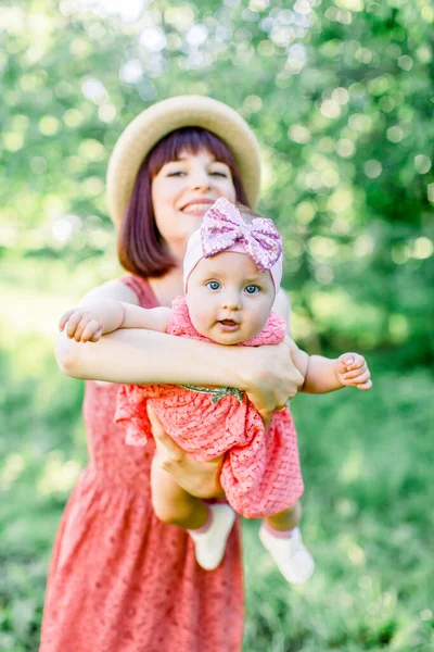 Hasır şapkalı güzel anne ve küçük kızı açık havada pembe bir elbise içinde. Aile görünüşü. Genç anne güneşli bir günde bebeği gökyüzüne kusar.. — Stok fotoğraf