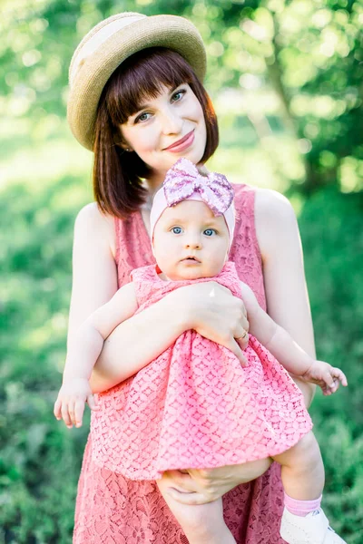 藁帽子の美しい母と彼女の小さな娘屋外の家族はピンクのドレスで見ています。幸せな家族の屋外肖像画。家族みたいに公園の肖像画の母親と赤ちゃん — ストック写真