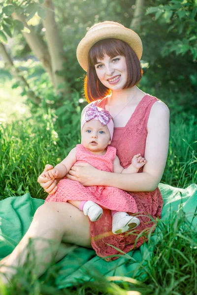春の晴れた日に緑の庭でピクニックをしている幸せな家族:美しい笑顔の母親は緑の草の上に座っていると彼女の足の上に彼女の小さな笑いの娘 — ストック写真