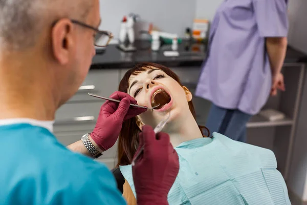 Jovem mulher bonita paciente na clínica odontológica com cheque dentário e tratamento, mulher com boca aberta, equipe de dentista profissional, dentista masculino sênior e sua assistente feminina — Fotografia de Stock