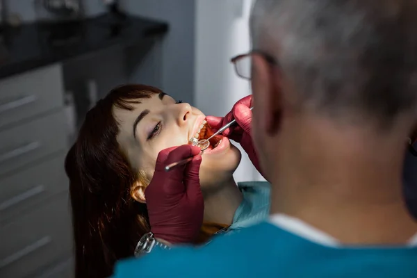 Oldalsó nézet a vezető férfi orvos fogorvos a fogorvos műtét, amelynek fogászati műtét fiatal nő beteg a modern fogászati klinikán. Koncentrálj a női arcra — Stock Fotó