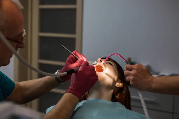 Fiatal beteg nő a fogorvosi rendelőben fogászati vizsgálat és kezelés fogászati eszközökkel idősebb férfi fogorvos és női asszisztens. Fogászati kezelés koncepciója — Stock Fotó