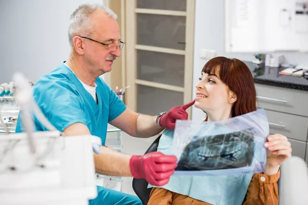 Smiling senior man dentist showing dental X-ray to pretty female patient, looking each other, preparing for dental treatment