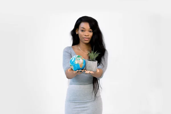 Ung afroamerikansk kvinna visar lite jord jordklotet i ena handen och lite grå kruka med växt i en annan, isolerad på vit bakgrund. Fokus på händer — Stockfoto