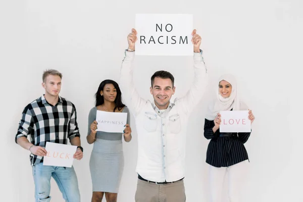 Stop racism, no racial discrimination of people. Young Caucasian man holds a poster with No racism text, while standing together with multiethnical friends with slogans on white background indoors — Stock Photo, Image