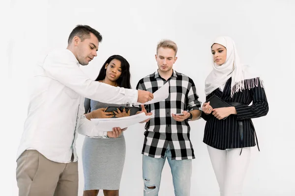 Vier befreundete multikulturelle College-Studenten, männlich und weiblich, stehen auf weißem Hintergrund, während ein kaukasischer Typ Papiere zum Studieren oder Neugründung für andere gibt — Stockfoto